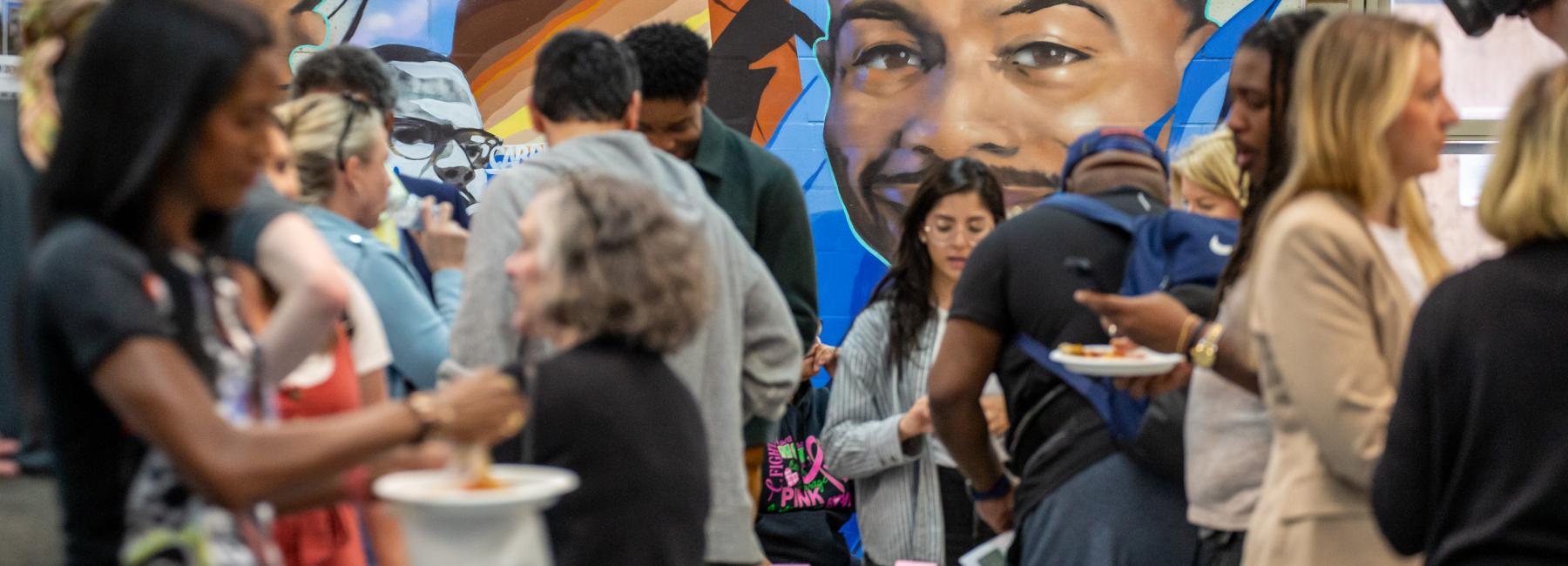A diverse group of people gathers in front of a vibrant mural during an event. The mural features the faces of African American individuals and abstract elements. The attendees are engaged in various activities, such as eating, talking, and looking at materials on a table. The setting appears to be an indoor community center or similar venue.