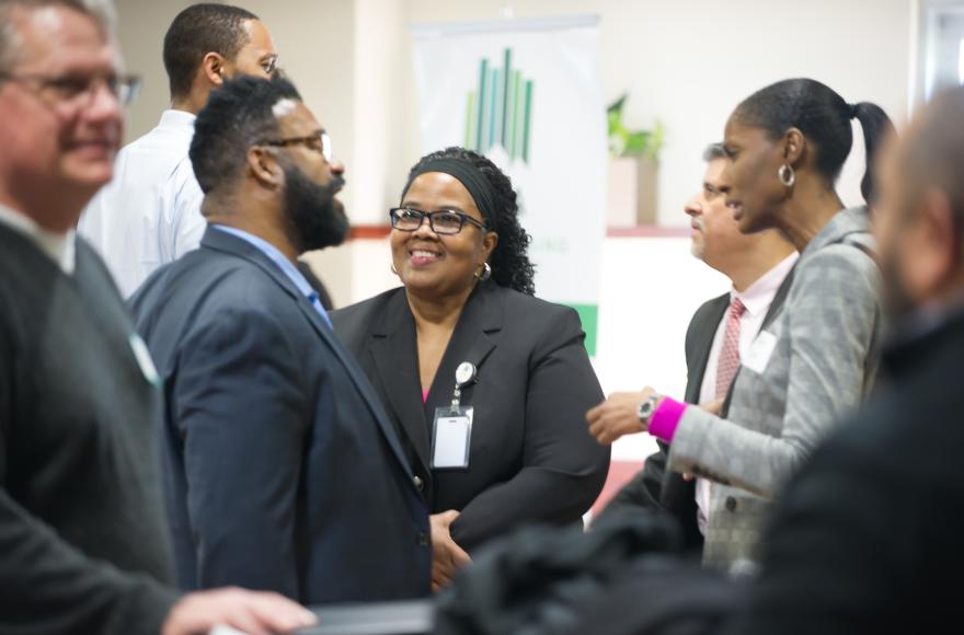 A group of professionals are gathered and engaging in conversation at a networking event or conference. One woman in the center is smiling, wearing a black suit and glasses. Others around her are also dressed in business attire, including suits and name tags. The setting appears to be a formal indoor event space.